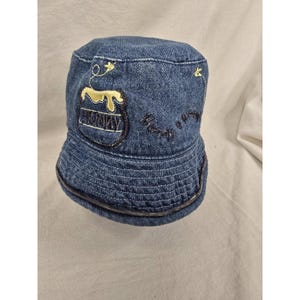 Puede incluir: Sombrero de pescador de mezclilla con bordado de tarro de miel y flores. El sombrero presenta un diseño de tarro de miel con la palabra "HONNY" bordada en la parte delantera. El sombrero es azul y tiene un borde cosido.
