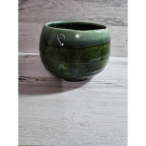 May include: A round, ceramic bowl with a glossy, green glaze. The bowl has a wide opening and a rounded shape. The color transitions from a lighter green at the top to a darker green towards the base. The bowl is likely handmade.