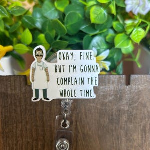 May include: A white and black badge reel featuring a cartoon character with the text "Okay, Fine. But I'm Gonna Complain The Whole Time." The badge reel has a clear plastic strap and a silver-colored metal clip.