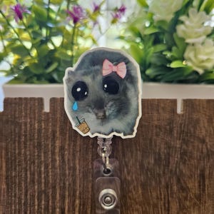 May include: A badge reel featuring a sad cartoon hamster with a pink bow, a single tear, and a small iced coffee. The hamster is gray and white with large black eyes. The reel is clear plastic.