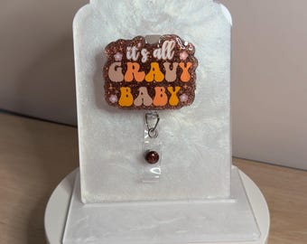 May include: A rectangular badge reel with a brown glitter finish featuring the text 'it's all GRAVY BABY' in white, orange, and yellow retro-style lettering, accented with small white flower icons and a clear plastic clip attachment.
