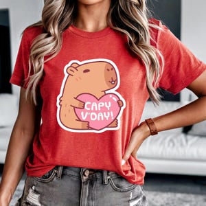 Könnte beinhalten: Rotes T-Shirt mit einem Cartoon-Capybara, das ein rosa Herz mit dem Text "CAPY V'DAY!" hält. Das Capybara ist hellbraun mit weißem Umriss. Das T-Shirt hat eine einfarbige rote Farbe.
