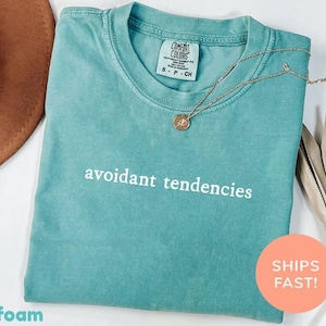 Könnte beinhalten: Türkisfarbenes T-Shirt mit dem weißen Schriftzug „avoidant tendencies“. Eine goldene Halskette mit einem kleinen Anhänger liegt auf dem Shirt. Ein brauner Hut und getrocknete Blätter sind im Hintergrund. Das Shirt hat ein Comfort Colors-Etikett.