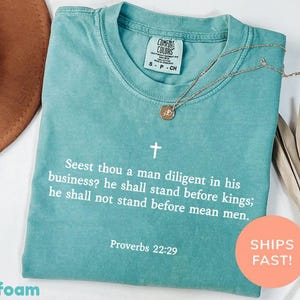 Può includere: Una t-shirt a maniche corte color verde schiuma di mare con una croce bianca e il testo: Seest thou a man diligent in his business? he shall stand before kings; he shall not stand before mean men. Proverbs 22:29. La maglietta ha un'etichetta Comfort Colors.