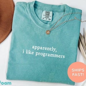 Könnte beinhalten: Meergrünes T-Shirt mit dem weißen Text "apparently, i like programmers". Das Shirt hat einen Rundhalsausschnitt und kurze Ärmel. Eine goldene Halskette mit Anhänger liegt auf dem Shirt. Ein brauner Hut und getrocknete Blätter sind im Hintergrund.
