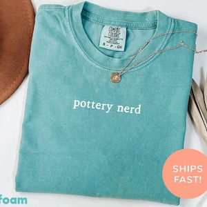 Puede incluir: Camiseta verde mar con el texto "pottery nerd" en blanco. Un collar dorado con un colgante descansa sobre la camiseta. La camiseta está doblada y se muestra con un sombrero marrón y hojas secas.