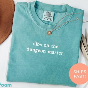 May include: Seafoam green t-shirt with the text "dibs on the dungeon master" in white. A gold necklace with a pendant rests on the shirt. The shirt has a Comfort Colors tag. A brown hat and dried leaves are in the background.