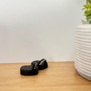 May include: Two black, heart-shaped earrings with a row of small, clear rhinestones on the top curve. The earrings are displayed on a light brown wooden surface, with a white background and a white, ribbed planter with greenery.