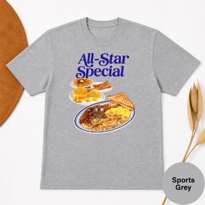 May include: Sports Grey t-shirt with a graphic of a breakfast plate, pancakes, and bacon. The text "All-Star Special" is printed in blue above the food illustration. The shirt is made of a soft material.