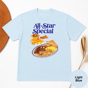 May include: Light blue t-shirt featuring the text "All-Star Special" in blue script above a graphic of a breakfast plate with pancakes, eggs, bacon, toast, and hash browns. The shirt is a light blue color.