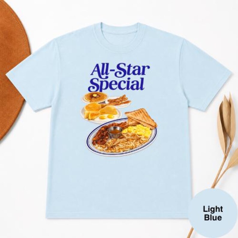 May include: Light blue t-shirt featuring the text "All-Star Special" in blue script above a graphic of a breakfast plate with pancakes, eggs, bacon, toast, and hash browns. The shirt is a light blue color.
