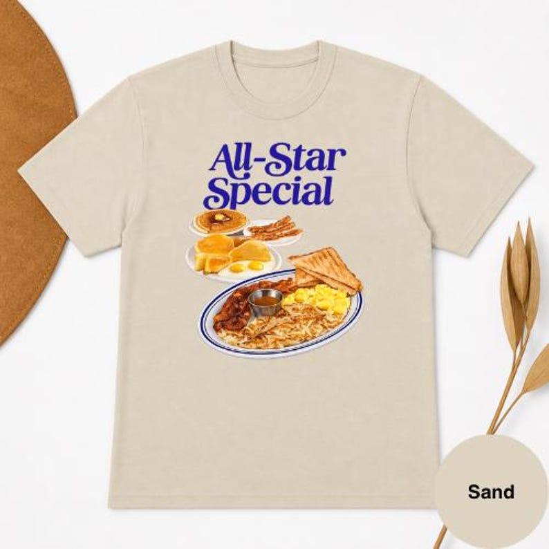 May include: Sand-colored t-shirt featuring the text "All-Star Special" in blue. The graphic depicts a breakfast plate with pancakes, eggs, bacon, toast, and hash browns. The shirt is made of a soft material.
