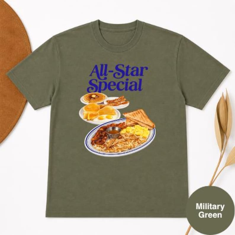 May include: Military green t-shirt featuring a graphic of a breakfast plate with pancakes, bacon, eggs, toast, and hash browns. The text "All-Star Special" is in blue script above the food illustration. The shirt is a solid color.