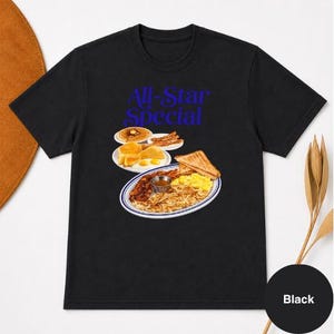 May include: Black t-shirt with a blue graphic that reads "All-Star Special." The graphic features a plate of breakfast food, including eggs, bacon, toast, and pancakes. A black circle with the word "Black" is in the bottom right corner.