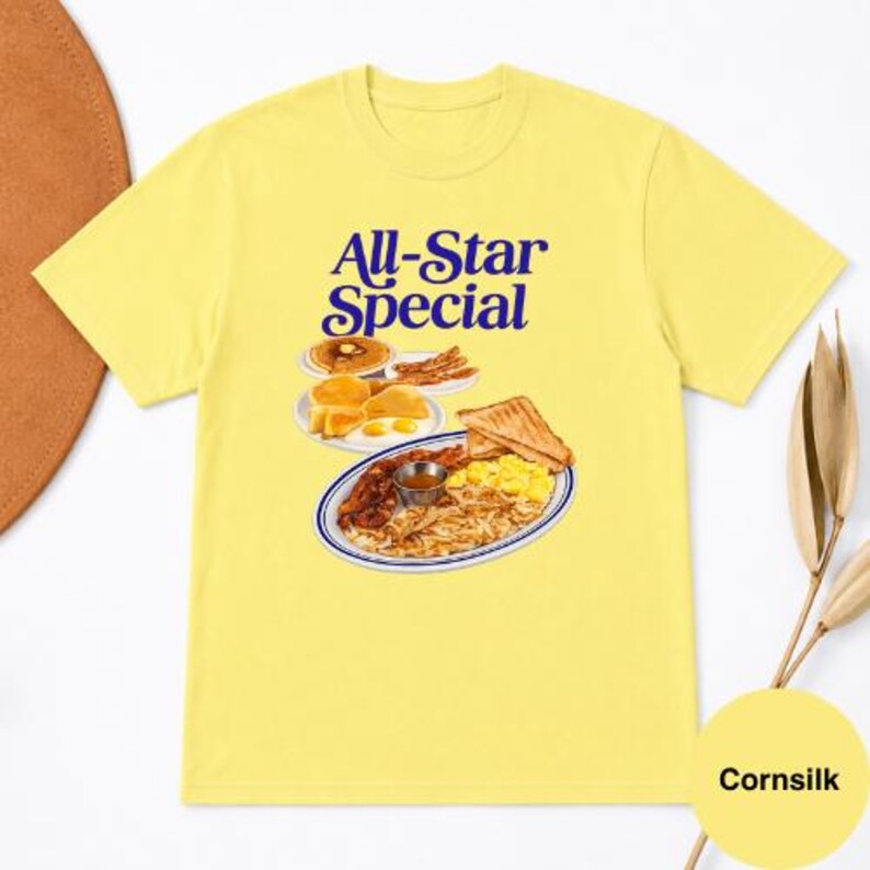 May include: A cornsilk yellow t-shirt with the text "All-Star Special" in blue script. The shirt features a graphic of a breakfast plate with pancakes, eggs, bacon, toast, and hash browns. The shirt is a casual wear item.