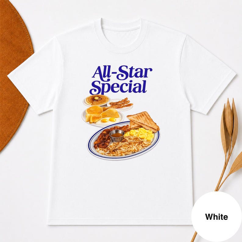 May include: White t-shirt featuring a colorful illustration of a breakfast platter with pancakes, eggs, bacon, and toast. The text "All-Star Special" is printed in blue script above the food image. The shirt is made of a soft material.