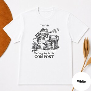 Op de afbeelding: Wit T-shirt met een zwart-wit illustratie van een kikker met een hooivork, naast een houten kist. De tekst op het shirt luidt: "That's it. You're going in the COMPOST."