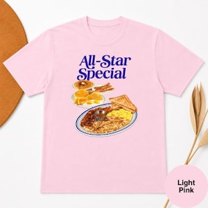 May include: Light pink t-shirt with the text "All-Star Special" in blue. The graphic features a breakfast plate with eggs, hash browns, and toast. Additional breakfast items include pancakes and bacon.
