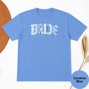 Puede incluir: Una camiseta de manga corta en color azul Carolina con la palabra BRIDE en letras de bloque en el pecho. Cada letra está rellena con un patrón de patchwork de varios diseños de tela azul y blanca con flores, cuadros y lunares.