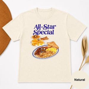 May include: Cream-colored t-shirt featuring a graphic of a breakfast plate with pancakes, bacon, eggs, and toast. The text "All-Star Special" is printed in blue above the food illustration. The shirt is made of a soft, natural fabric.