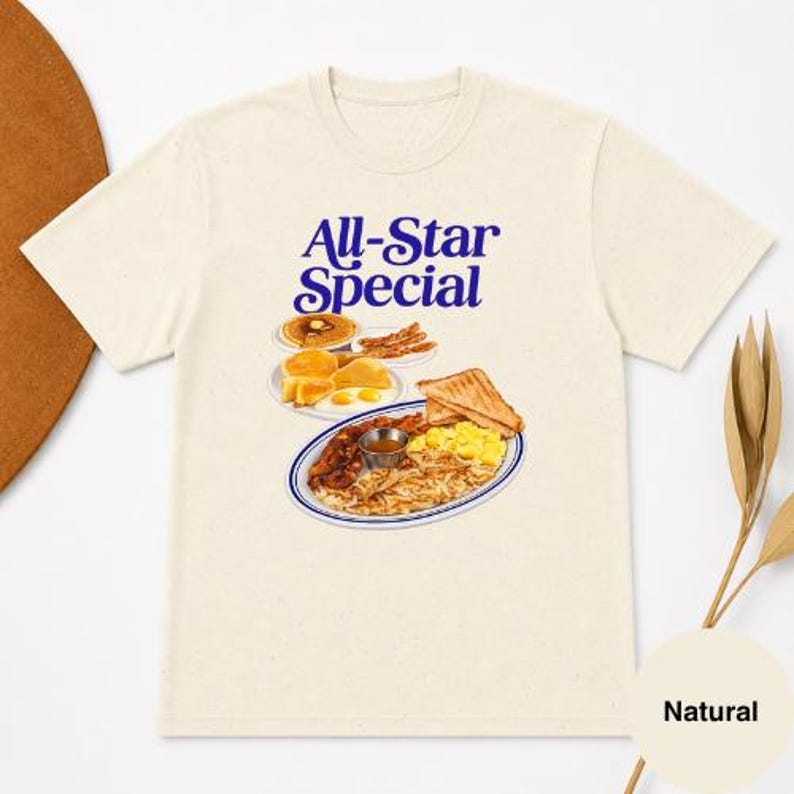May include: Cream-colored t-shirt featuring a graphic of a breakfast plate with pancakes, bacon, eggs, and toast. The text "All-Star Special" is printed in blue above the food illustration. The shirt is made of a soft, natural fabric.