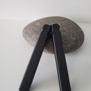 May include: A black butterfly knife, open, resting on a gray stone. The knife's handles are rectangular and connected at the top. The background is a plain white surface.
