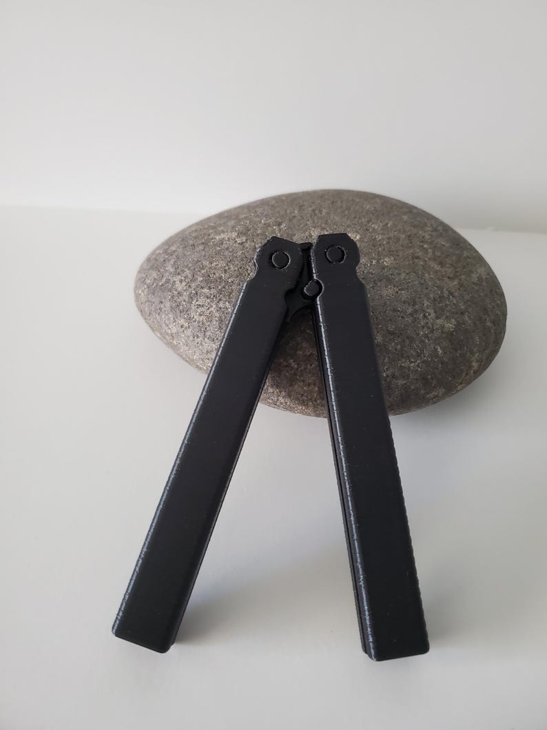 May include: A black butterfly knife, open, resting on a gray stone. The knife's handles are rectangular and connected at the top. The background is a plain white surface.