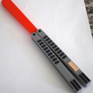 May include: A gray and black butterfly knife with an orange handle. The knife features a series of rectangular cutouts along the handle and a gold-colored accent near the base.