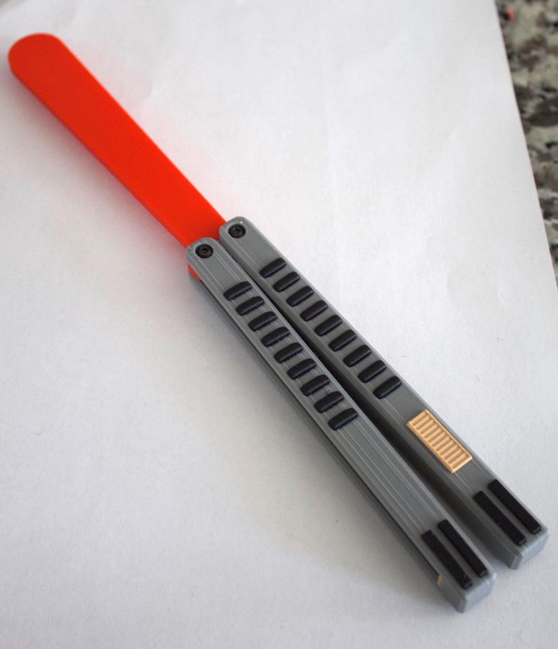 May include: A gray and black butterfly knife with an orange handle. The knife features a series of rectangular cutouts along the handle and a gold-colored accent near the base.