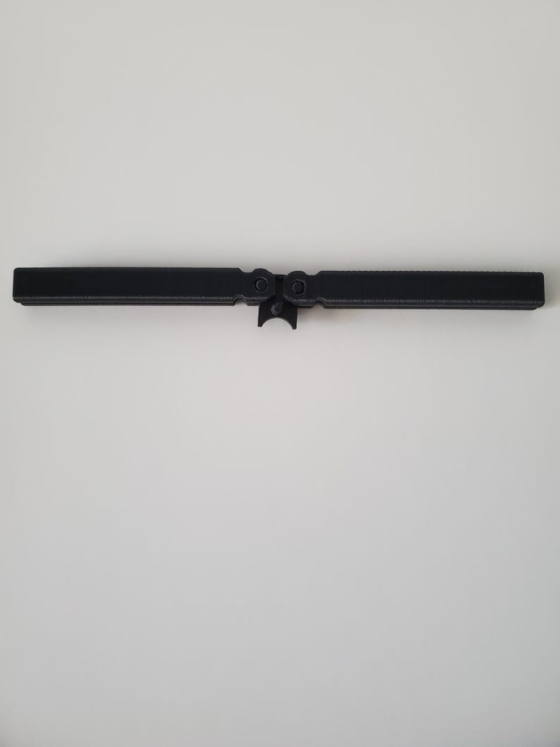 May include: A black, hinged metal object, possibly a tool or hardware component, is centered against a white background. The object has a rectangular shape with a central hinge and circular features on either side.