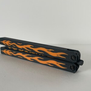 May include: A closed butterfly knife trainer featuring a matte black finish on the handles with a vibrant orange flame pattern printed along the sides. The folding tool includes two pivot screws and a small latch mechanism at the base.