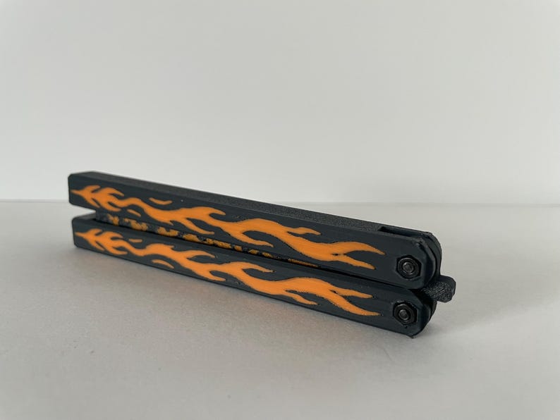 May include: A closed butterfly knife trainer featuring a matte black finish on the handles with a vibrant orange flame pattern printed along the sides. The folding tool includes two pivot screws and a small latch mechanism at the base.