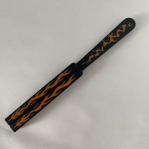 May include: A black butterfly knife trainer featuring orange flame patterns on the two handle segments and an orange jagged lightning or crack design on the single blade. The tool has a pivot mechanism connecting the handles to the blade.