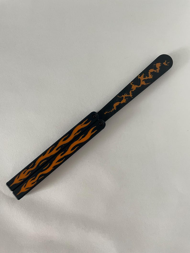 May include: A black butterfly knife trainer featuring orange flame patterns on the two handle segments and an orange jagged lightning or crack design on the single blade. The tool has a pivot mechanism connecting the handles to the blade.