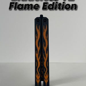 May include: A black and orange Bladeless V2 Flame Edition butterfly knife trainer. The closed tool features a dark handle with a prominent orange flame pattern running vertically along the center. The text Bladeless V2 Flame Edition is visible.