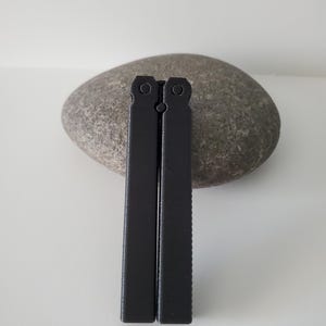 May include: Two black metal tools, possibly for a specific craft or hobby, are displayed vertically against a gray stone. The tools have a rectangular shape with a textured edge on one side. The background is a plain white surface.
