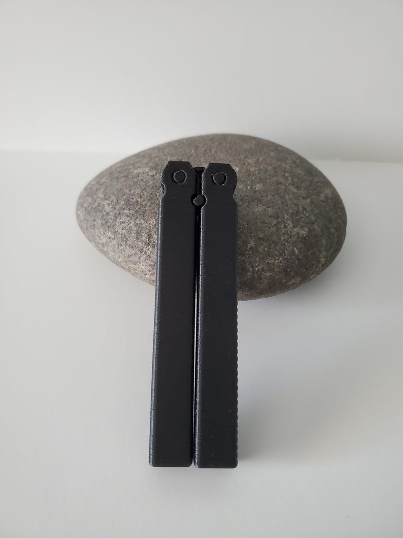 May include: Two black metal tools, possibly for a specific craft or hobby, are displayed vertically against a gray stone. The tools have a rectangular shape with a textured edge on one side. The background is a plain white surface.