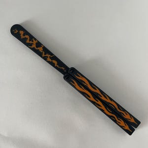 May include: A black butterfly knife trainer featuring a long blade with a textured orange lightning bolt pattern and two handles decorated with orange flame graphics. The folding tool is shown in a closed position with visible pivot screws.