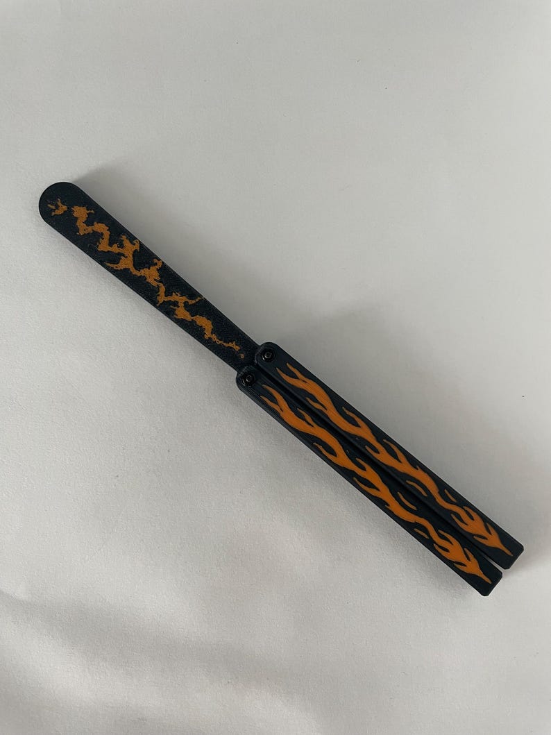 May include: A black butterfly knife trainer featuring a long blade with a textured orange lightning bolt pattern and two handles decorated with orange flame graphics. The folding tool is shown in a closed position with visible pivot screws.