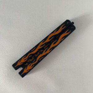 May include: A closed butterfly knife featuring two black handles with a vibrant orange flame pattern printed along the length of each side. The metal handles are connected by a pivot mechanism at the top, displaying a sleek and uniform design.