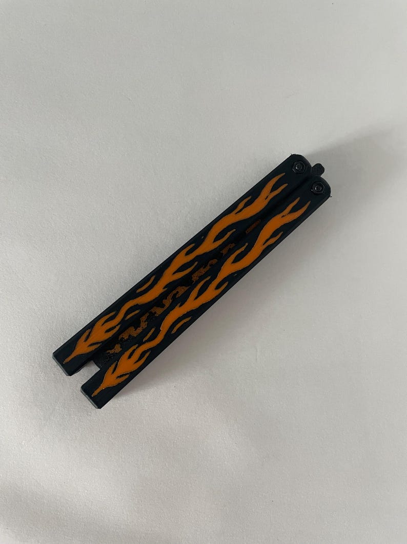 May include: A closed butterfly knife featuring two black handles with a vibrant orange flame pattern printed along the length of each side. The metal handles are connected by a pivot mechanism at the top, displaying a sleek and uniform design.