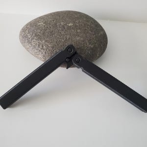 May include: A black butterfly knife, open, with a matte finish, lies on a white surface. The knife is positioned in front of a large, gray, oval-shaped stone. The knife's handles are extended, showcasing its design.