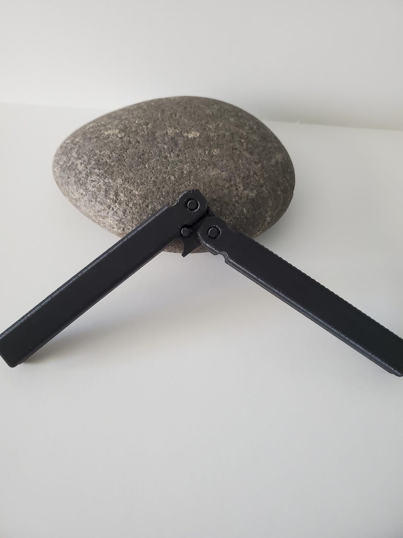 May include: A black butterfly knife, open, with a matte finish, lies on a white surface. The knife is positioned in front of a large, gray, oval-shaped stone. The knife's handles are extended, showcasing its design.