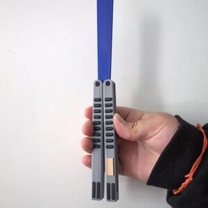 May include: A gray and blue butterfly knife, held against a white wall. The knife features a blue blade and gray handles with a series of vertical grooves. The handle has a small, rectangular, tan-colored accent. The knife is held in a hand.