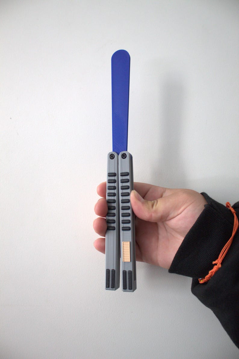 May include: A gray and blue butterfly knife, held against a white wall. The knife features a blue blade and gray handles with a series of vertical grooves. The handle has a small, rectangular, tan-colored accent. The knife is held in a hand.