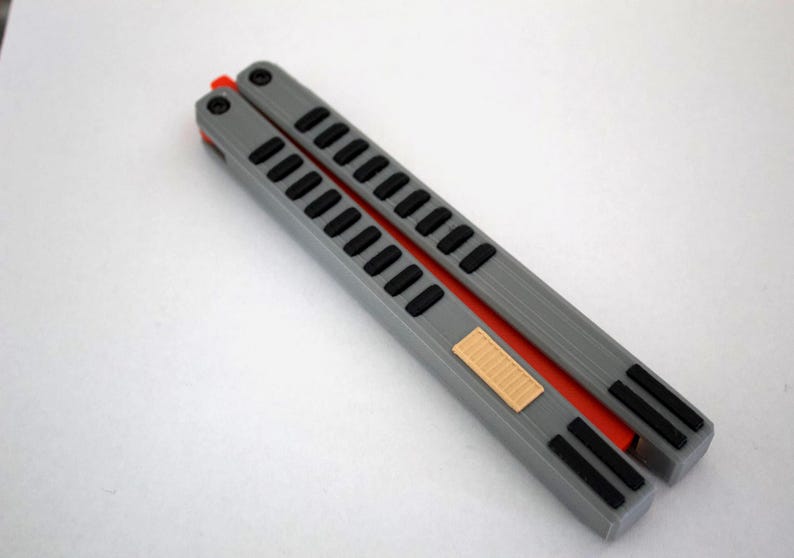 May include: A gray butterfly knife with black accents and an orange spine. The handle features a series of rectangular cutouts and a small, tan rectangular detail. The knife is designed for flipping and training.