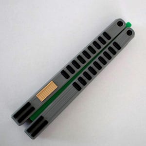 May include: A gray butterfly knife with black accents and a green spine. The handle features a series of rectangular cutouts and a tan-colored latch. The knife is displayed on a white surface.