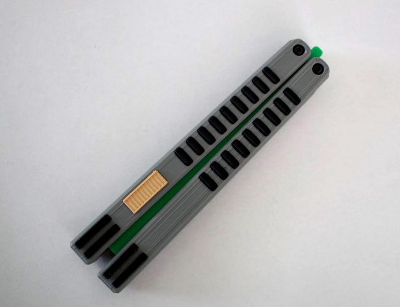 May include: A gray butterfly knife with black accents and a green spine. The handle features a series of rectangular cutouts and a tan-colored latch. The knife is displayed on a white surface.