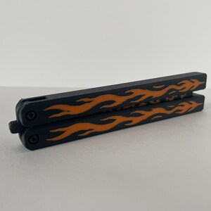 May include: A closed butterfly knife featuring a matte black finish on the handles with a prominent orange flame pattern printed along the length of each side. The two handles are secured together by a small black latch at the pivot end.