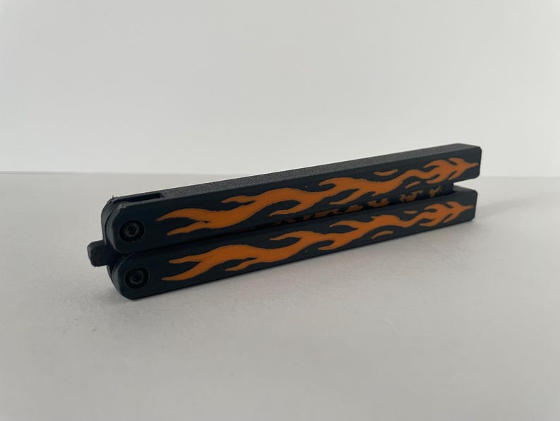 May include: A closed butterfly knife featuring a matte black finish on the handles with a prominent orange flame pattern printed along the length of each side. The two handles are secured together by a small black latch at the pivot end.